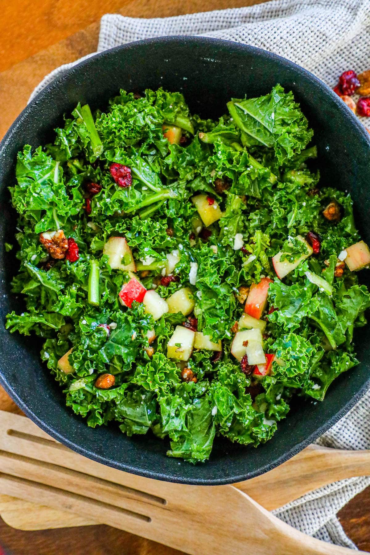 chopped kale, craisins, chopped apples, walnuts, and feta cheese crumbles tossed in dressing in a black bowl on a table next to wooden spoons