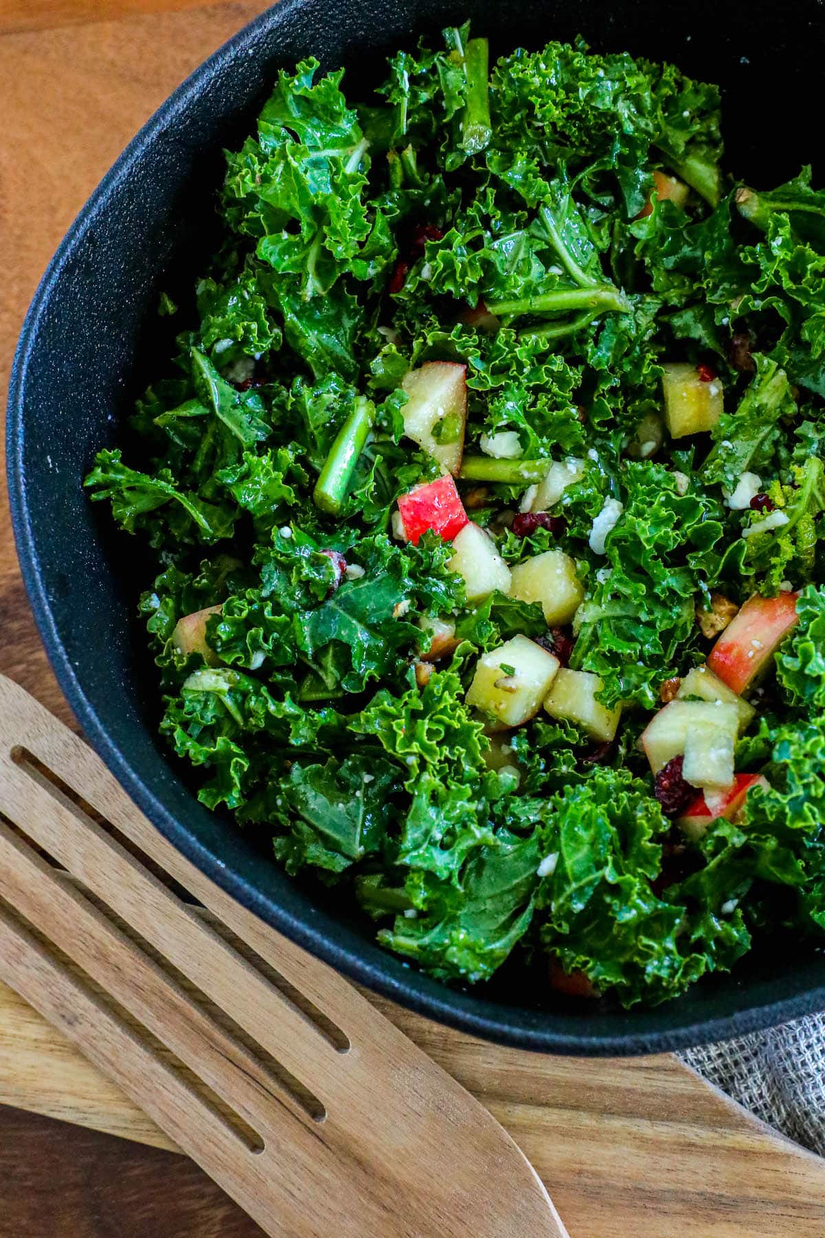 chopped curly kale, craisins, chopped apples, walnuts, and feta cheese crumbles tossed in dressing in a black bowl on a table next to wooden spoons