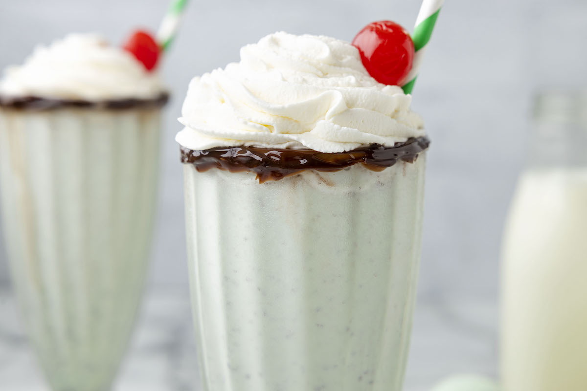 mint milkshake in a tall glass cup with whipped cream and a cherry on top, and a straw on the side