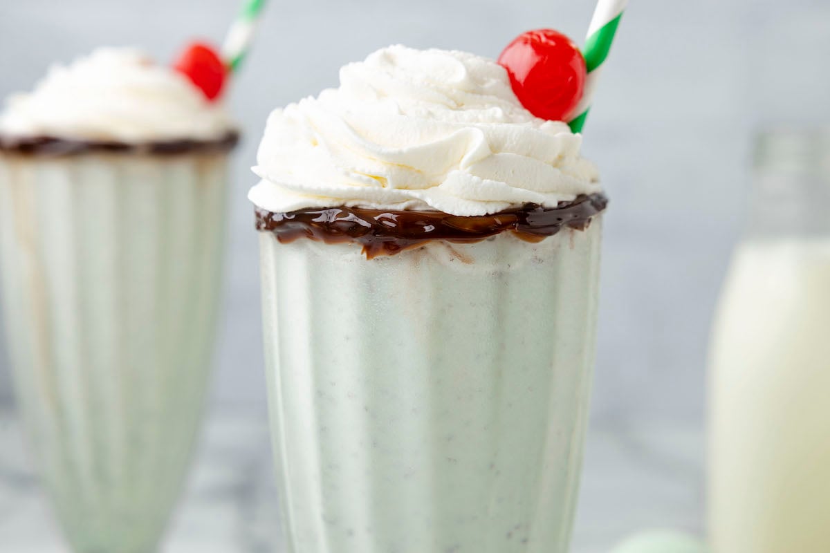 mint milkshake in a tall glass cup with whipped cream and a cherry on top, and a straw on the side