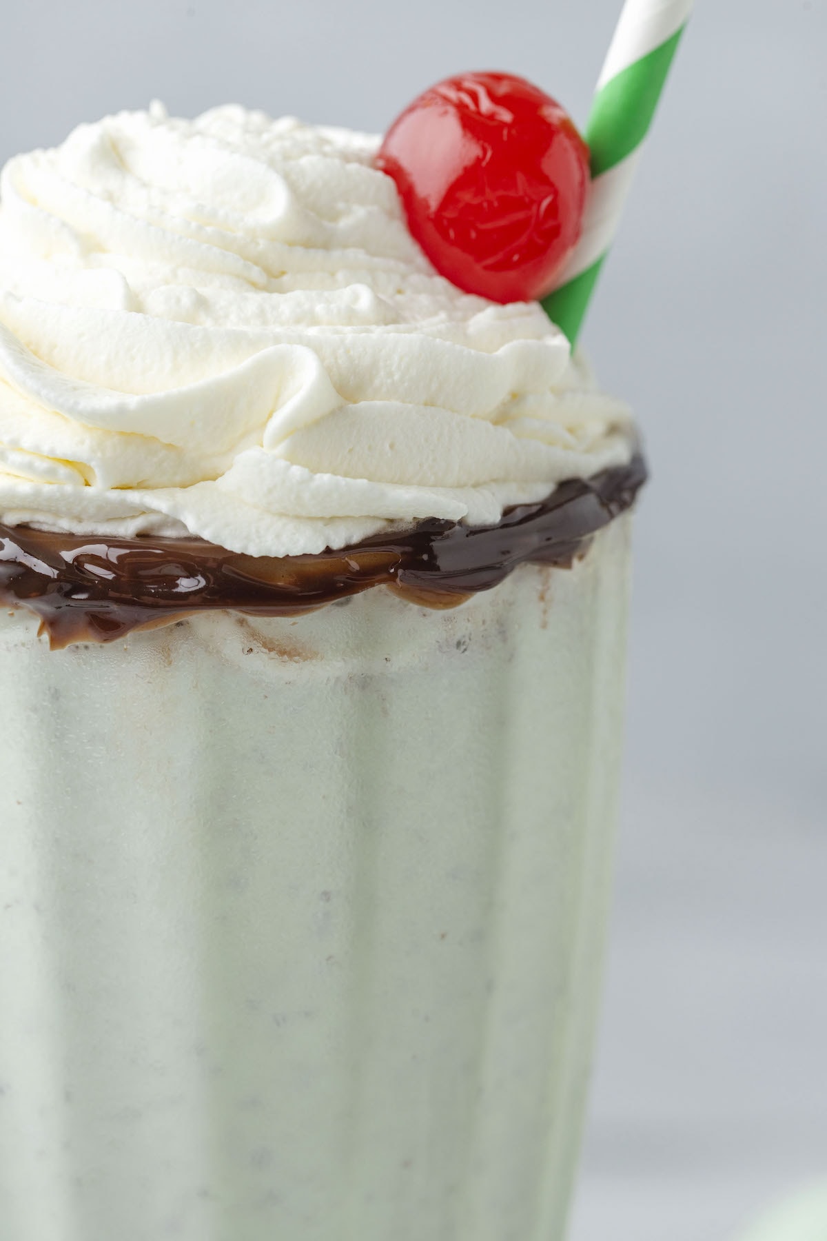 mint milkshake in a tall glass cup rimmed with chocolate with whipped cream and a cherry on top, and a straw on the side