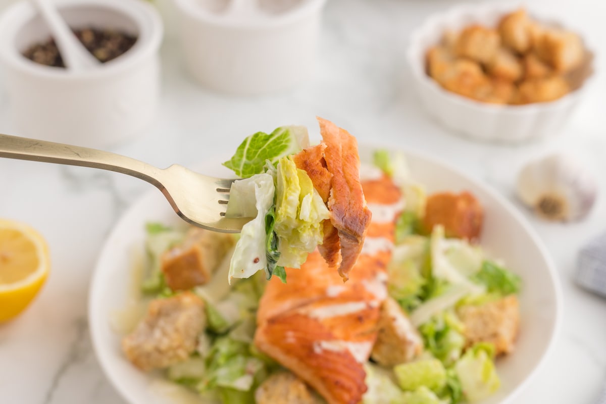 Salmon and salad on a fork in front of caesar salad with croutons in a white plate with a filet of salmon on top