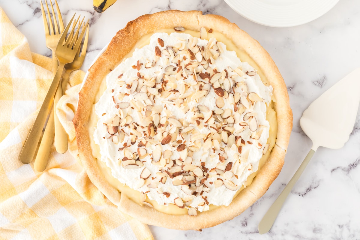 slice of banana cream pie on a plate with almonds on top and bananas on a table with a pie knife next to it 