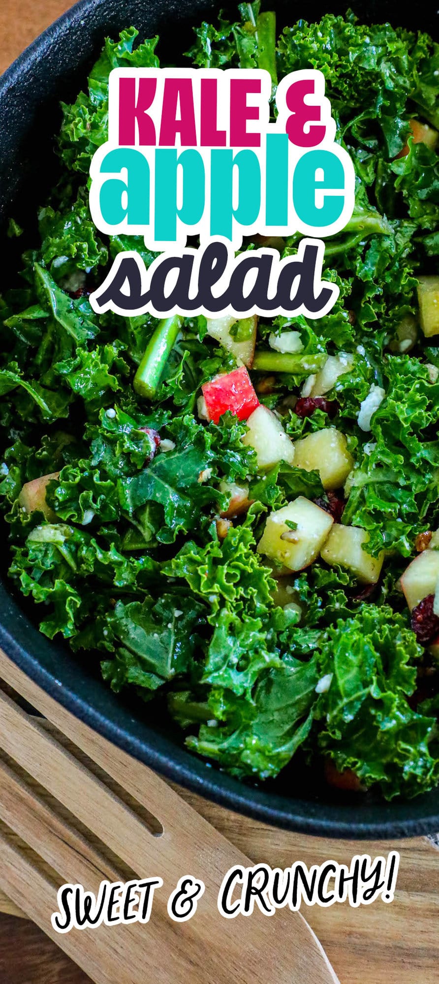 chopped green kale, craisins, chopped apples, walnuts, and feta cheese crumbles tossed in dressing in a black bowl on a table next to wooden spoons