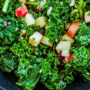 bright green kale, craisins, chopped apples, walnuts, and feta cheese crumbles tossed in dressing in a black bowl on a table next to wooden spoons