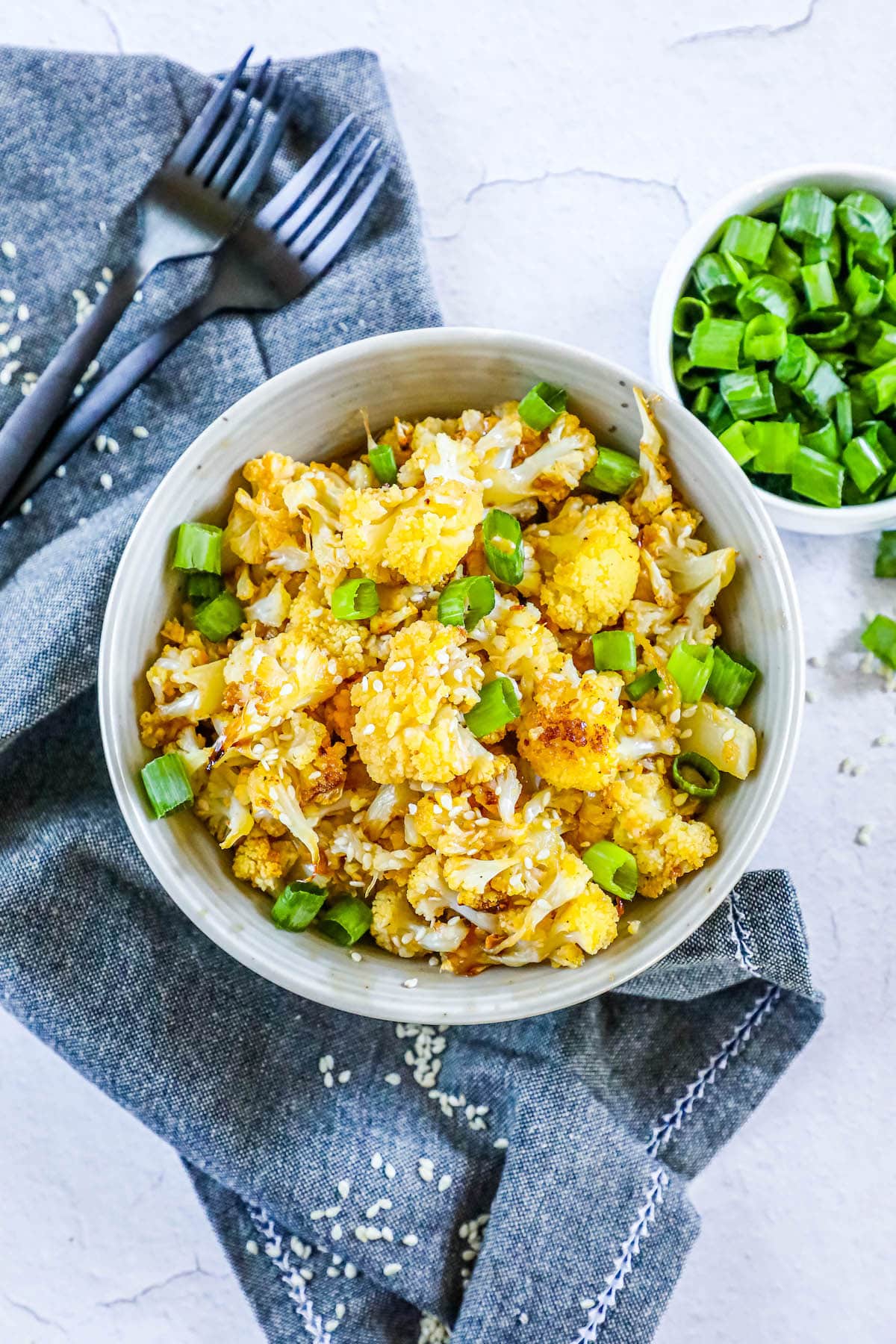 baked cauliflower with sesame seeds, sliced green onions, and seasoning in a white bowl on a table with forks next to the bowl 