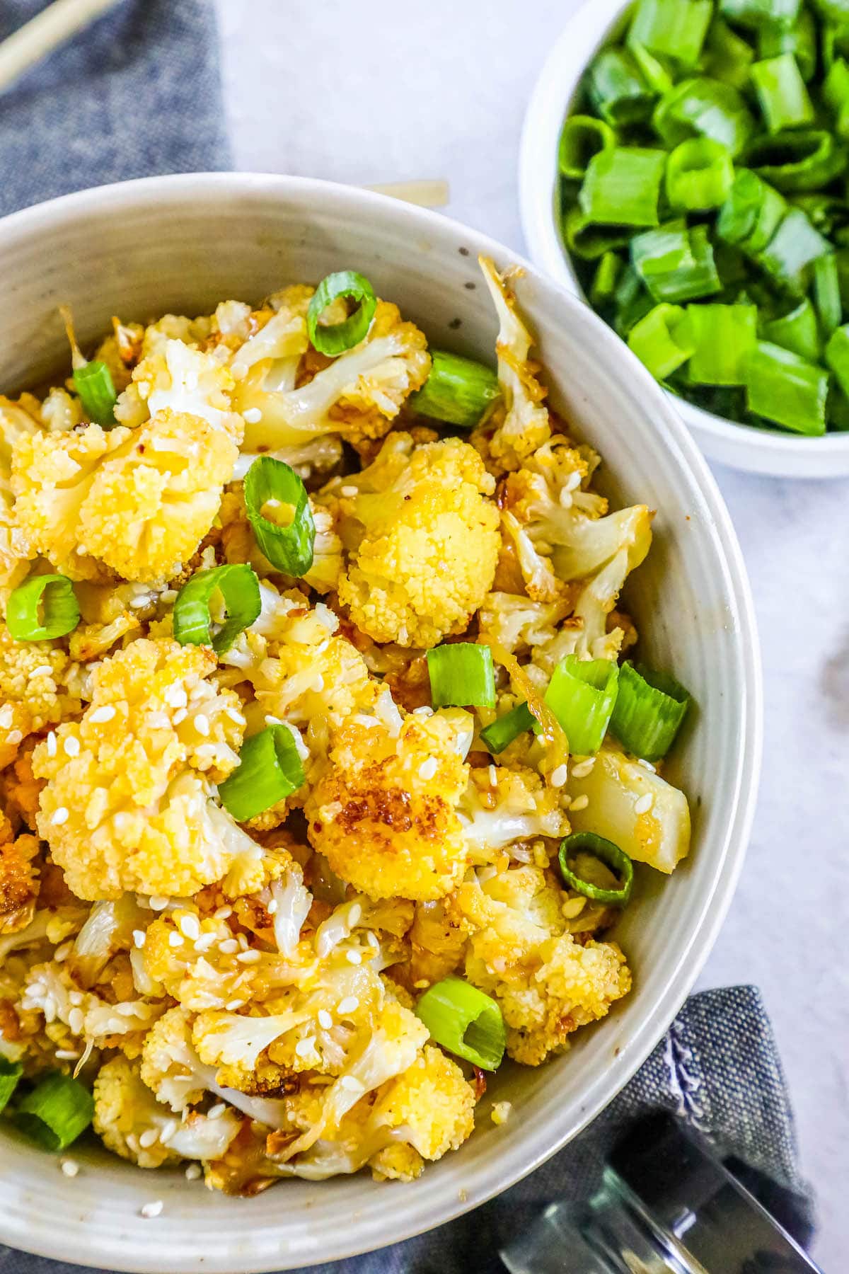 baked cauliflower with sesame seeds, sliced green onions, and seasoning in a white bowl on a table