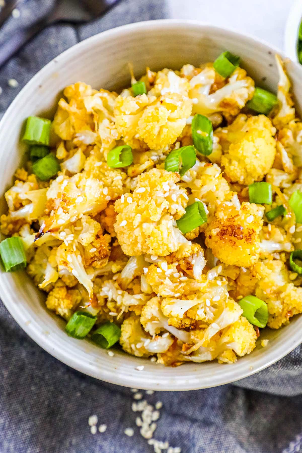 baked cauliflower with sesame seeds, sliced green onions, and seasoning in a white bowl on a table