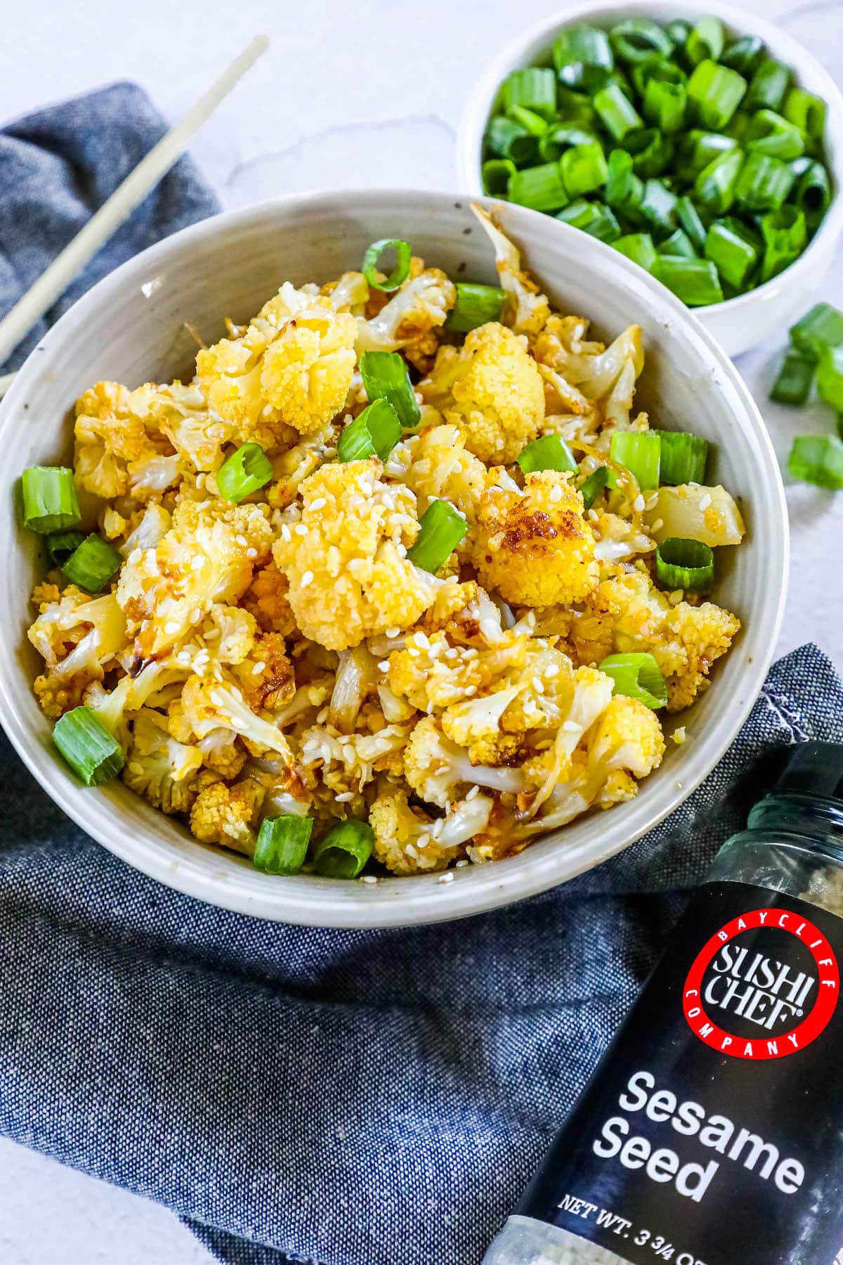 baked cauliflower with sesame seeds, sliced green onions, and seasoning in a white bowl on a table