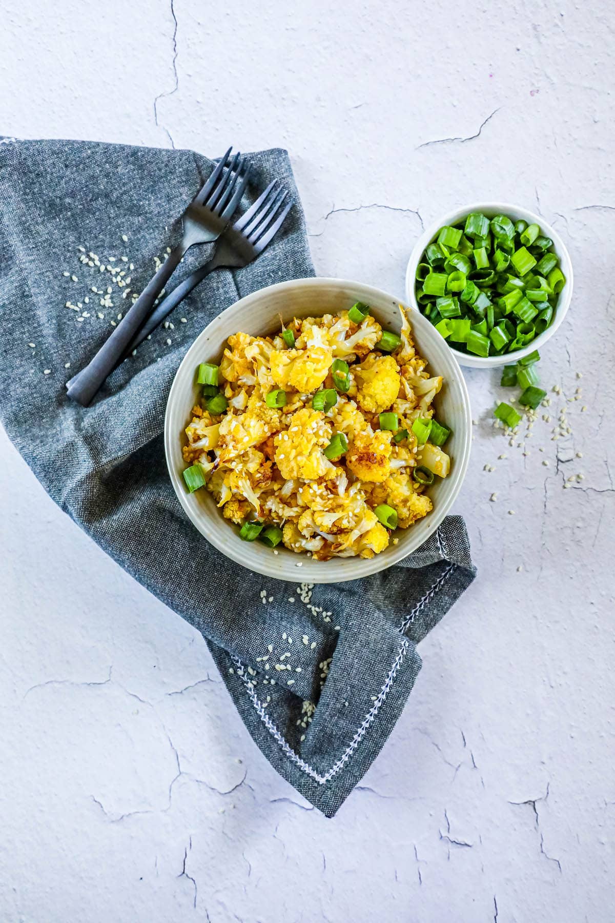 baked cauliflower with sesame seeds, sliced green onions, and seasoning in a white bowl on a table