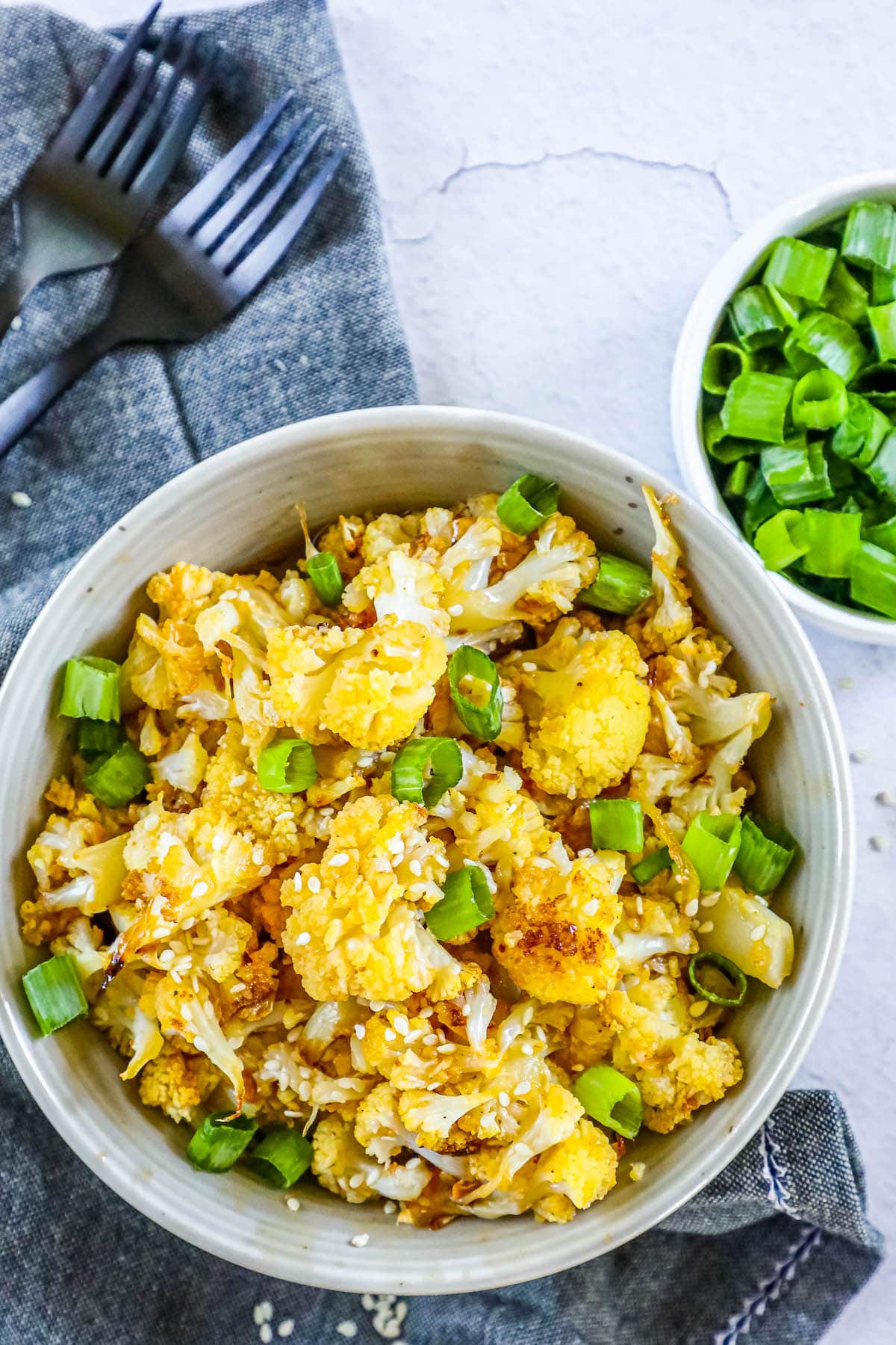 baked cauliflower with sesame seeds, sliced green onions, and seasoning in a white bowl on a table