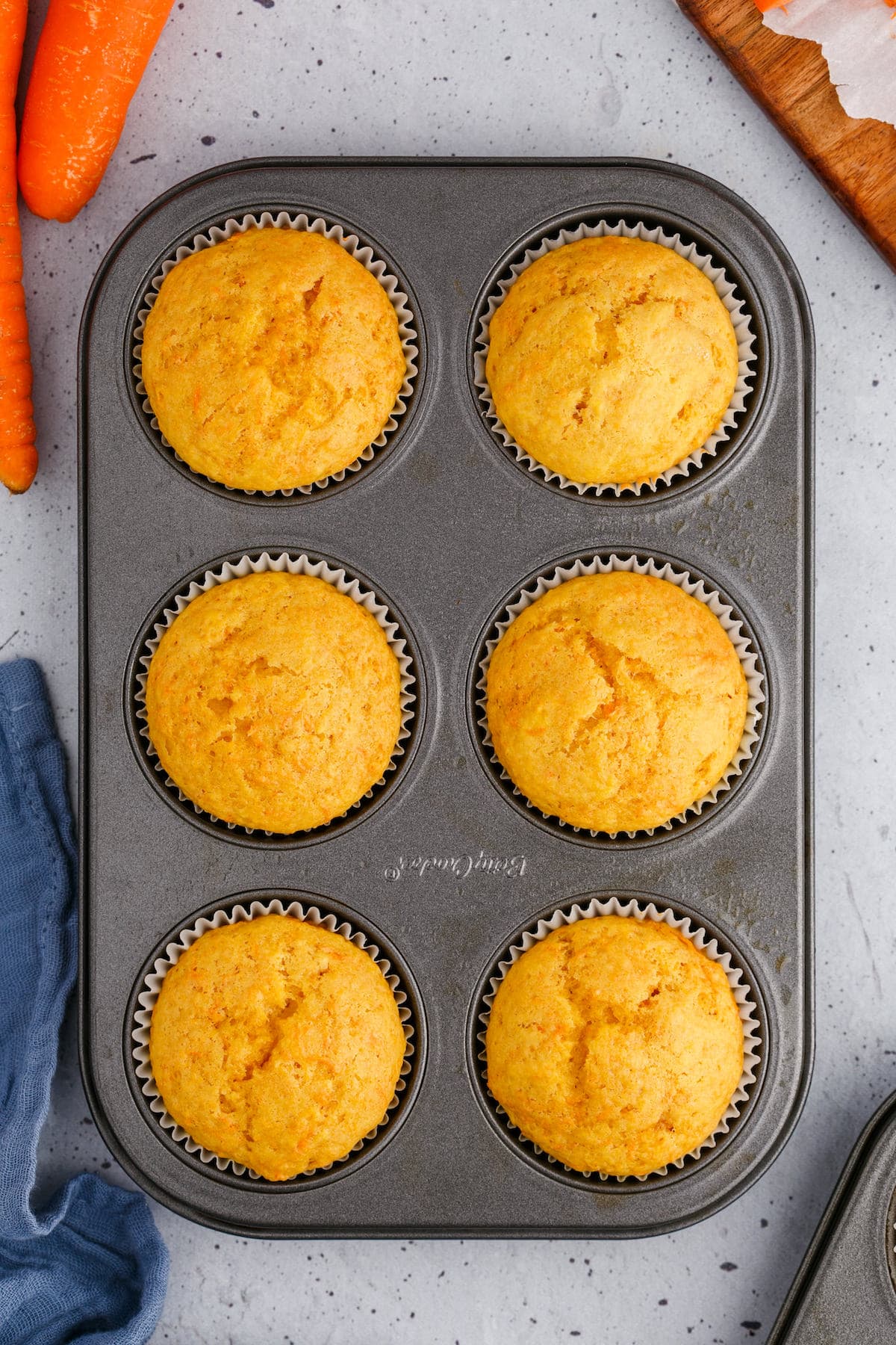 6 baked carrot muffins in a muffin tin on a table next to fresh carrots
