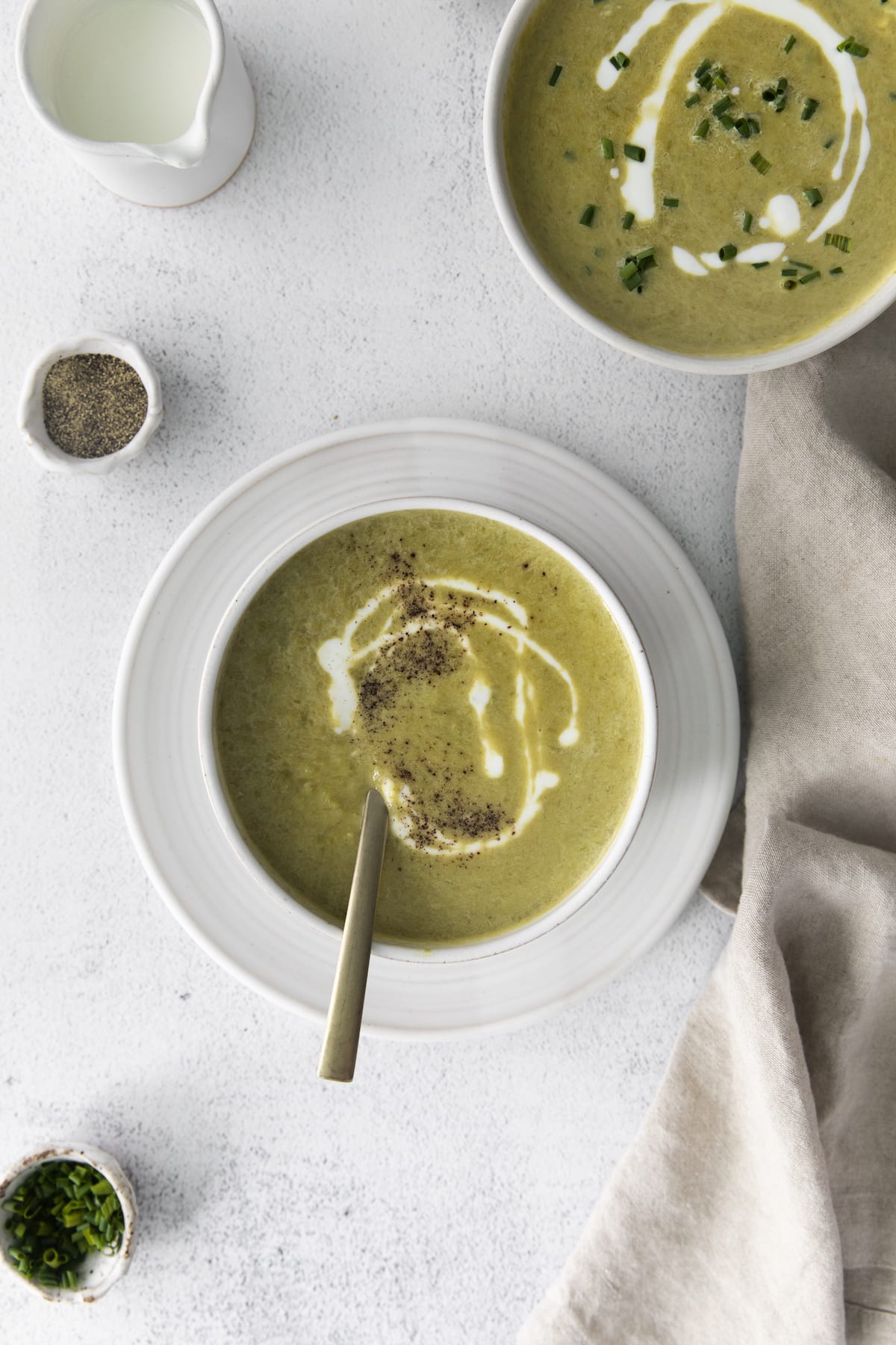 a spoon in creamy asparagus soup in a white bowl with a swirl of cream on top and lots of fresh cracked pepper 