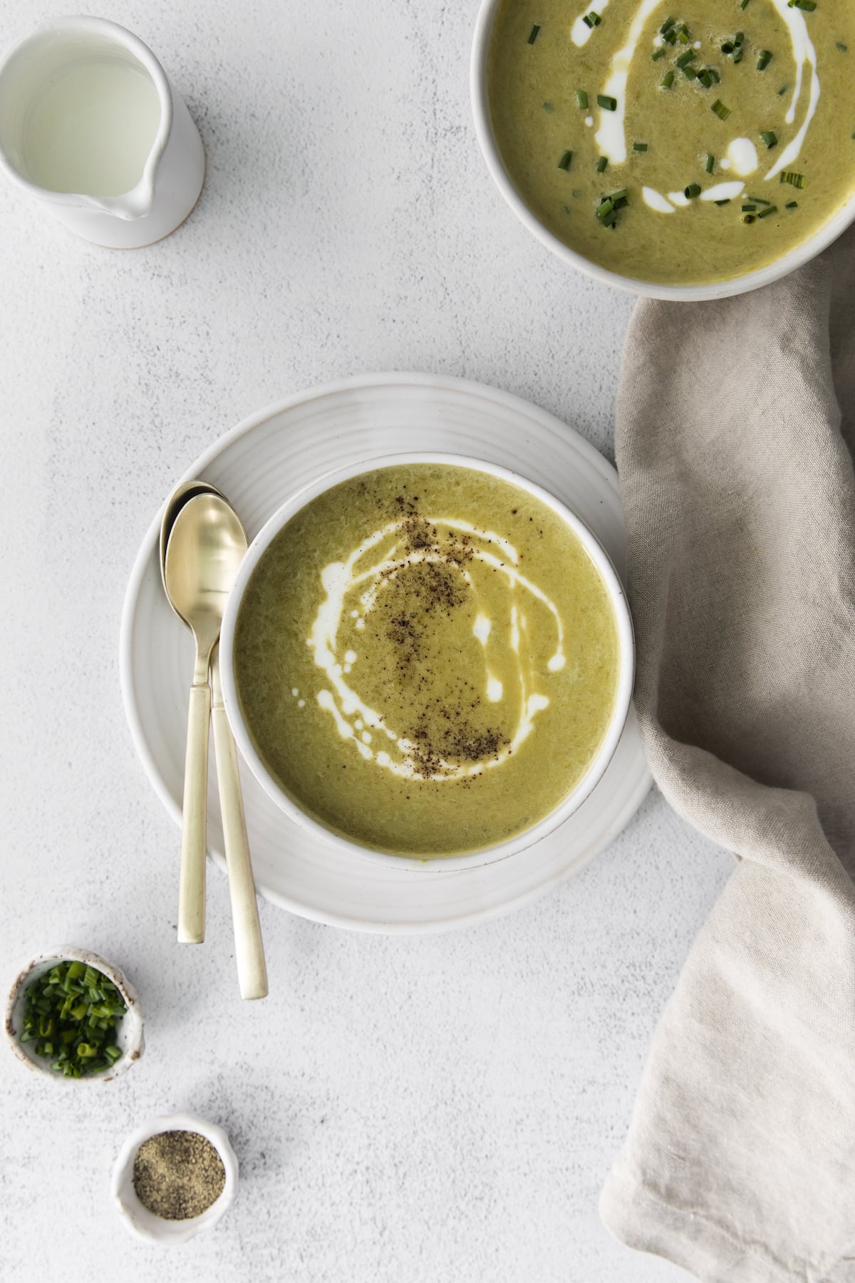creamy asparagus soup in a white bowl with a swirl of cream on top and lots of fresh cracked pepper next to a bowl of soup with chopped chives and cream on top 