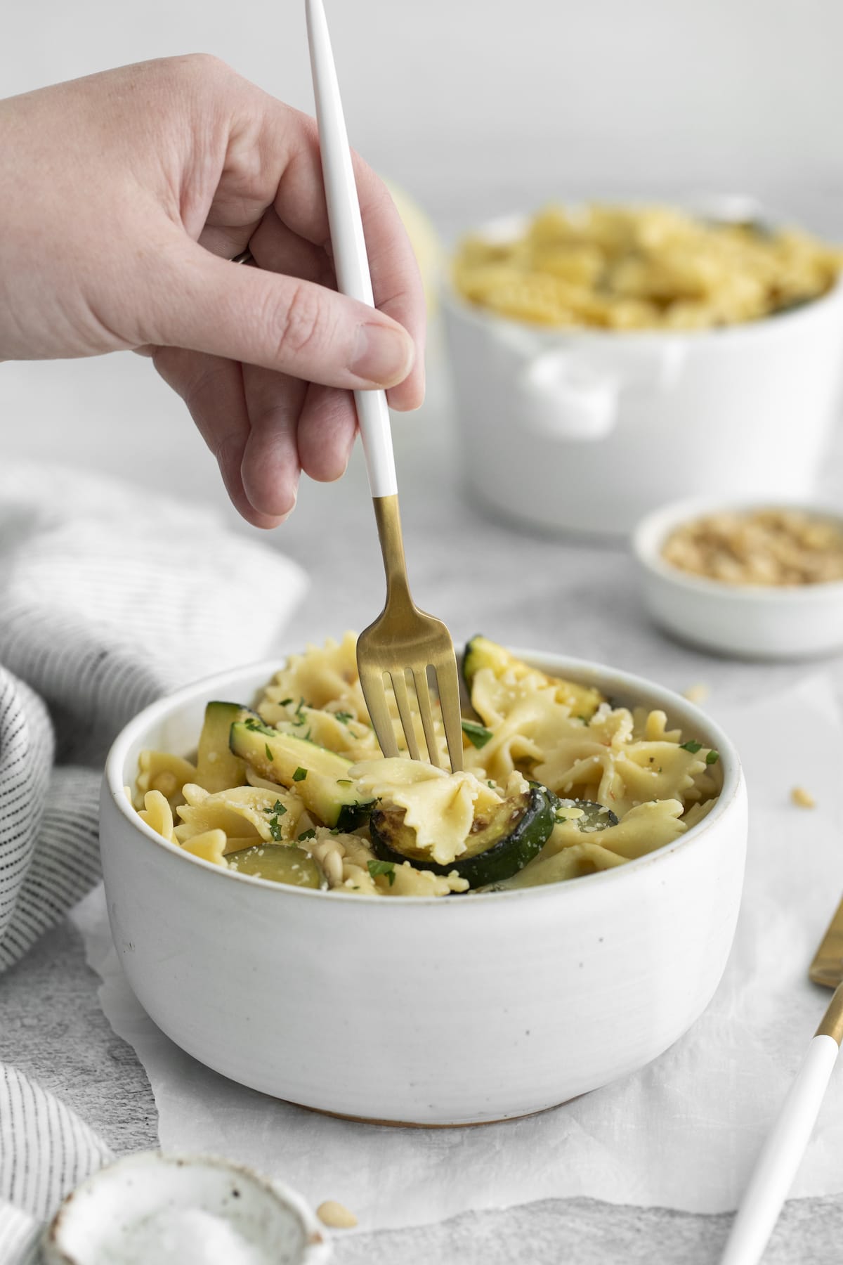 a hand holding a fork scooping up pasta with zucchini and herbs in a white bowl 