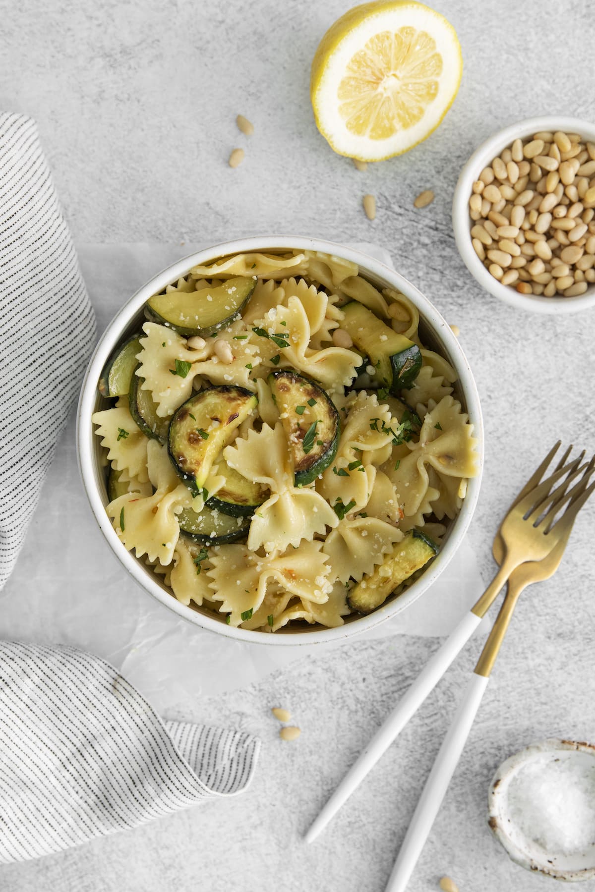 bowtie pasta with browned zuccini slices, parmesan cheese, pine nuts, and herbs in a bowl on a table next to a fork and a lemon cut in half