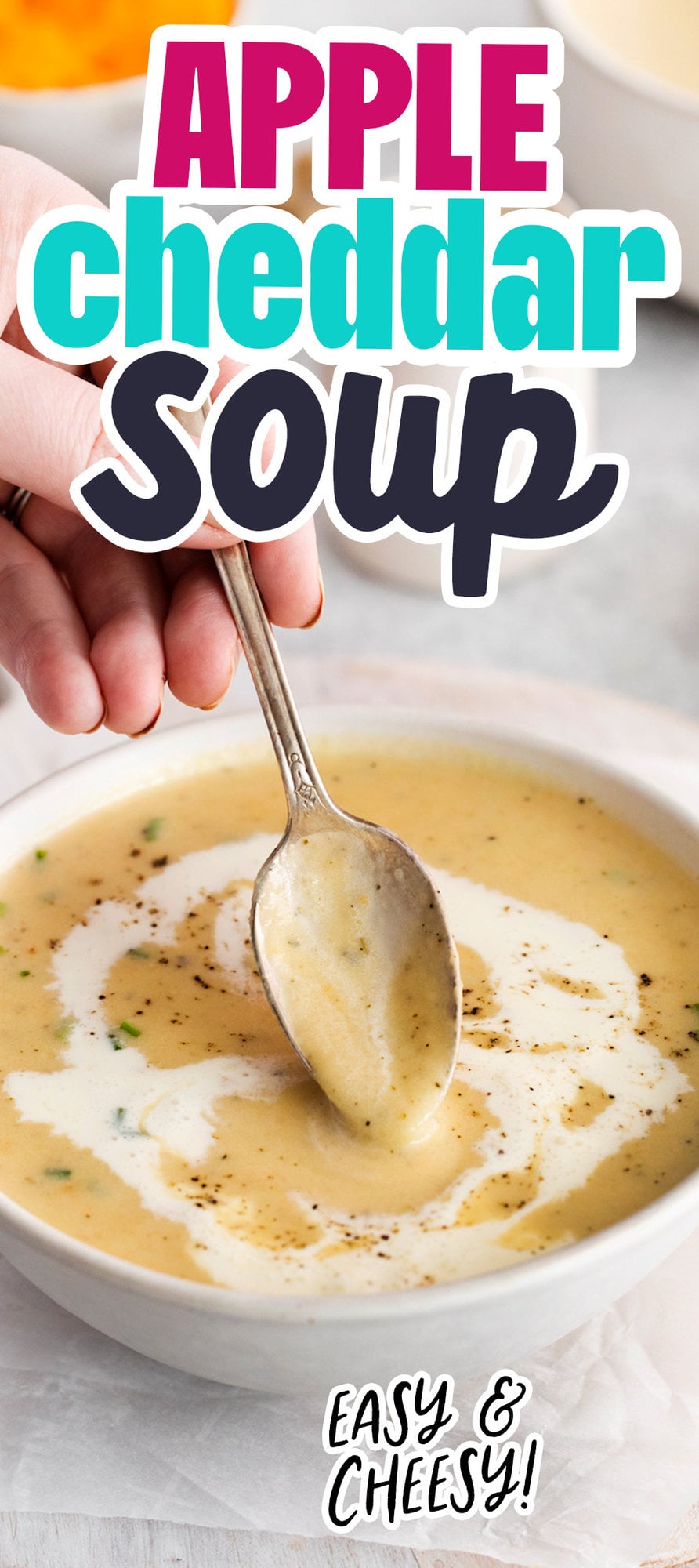 a hand holding a spoon dipping into a bowl of cheddar soup with a swirl of cream and crack of pepper on top
