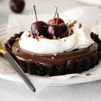 chocolate ganache tart in an oreo crust with whipped cream and fresh cherries on top on a white plate with a fork next to it