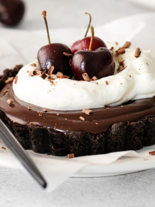chocolate ganache tart in an oreo crust with whipped cream and fresh cherries on top on a white plate with a fork next to it