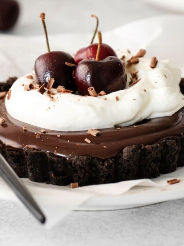 chocolate ganache tart in an oreo crust with whipped cream and fresh cherries on top on a white plate with a fork next to it