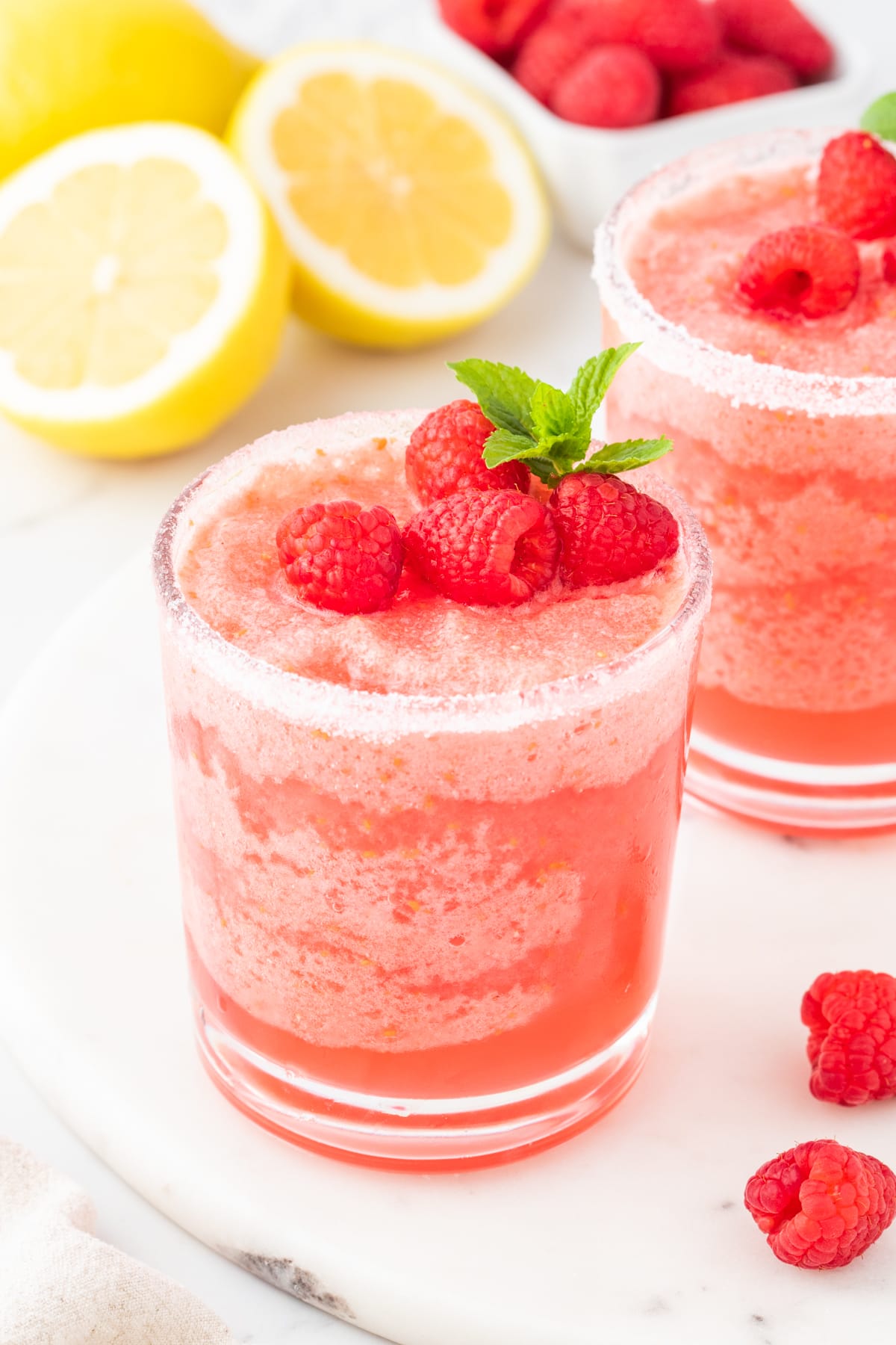 frozen pink lemonade in a sugar rimmed glass with raspberries and a sprig of mint on top next to another glass of lemonade