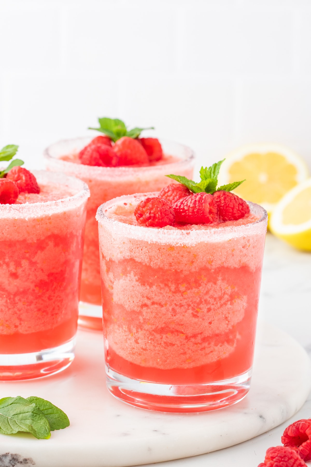 three frozen pink lemonades in  sugar rimmed glasses with raspberries and a sprig of mint on top