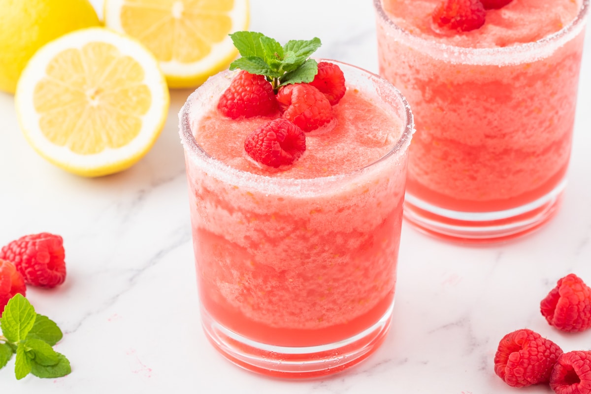 frozen pink lemonade in a sugar rimmed glass with raspberries and a sprig of mint on top next to another glass of raspberry lemonade on a table with raspberries and lemons on it 