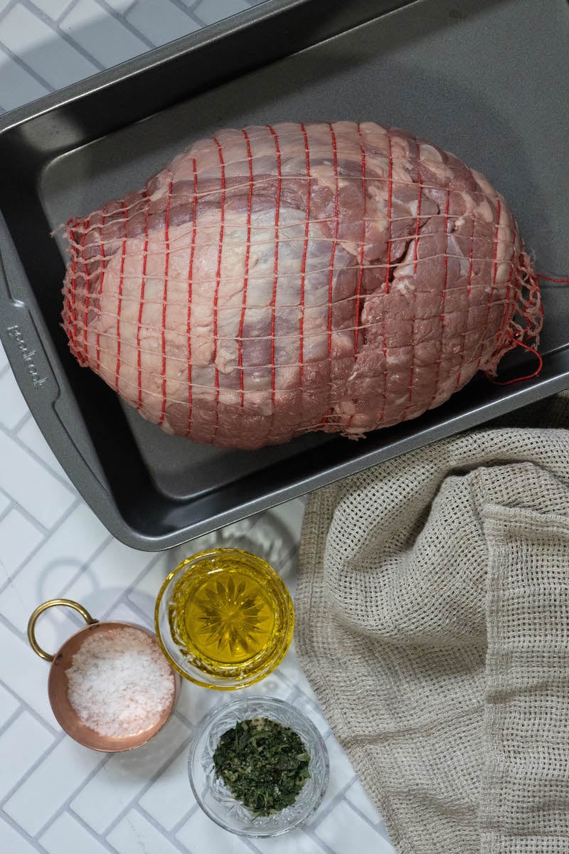 raw boneless leg of lamb in netting, olive oil, salt, and herbs on a table