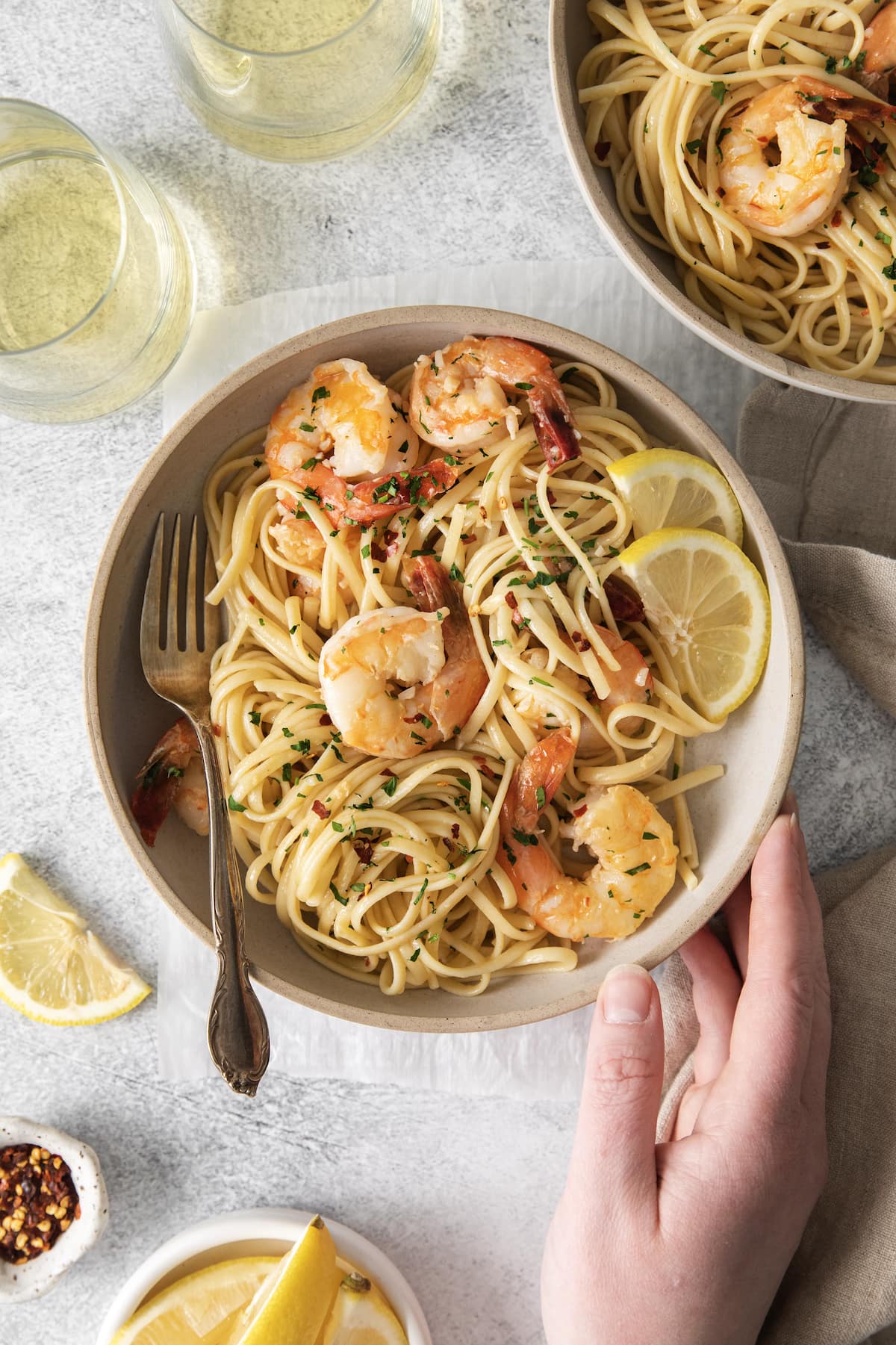 a hand touching a bowl with shrimp pasta with linguine, red pepper flakes, herbs, and lemon slices