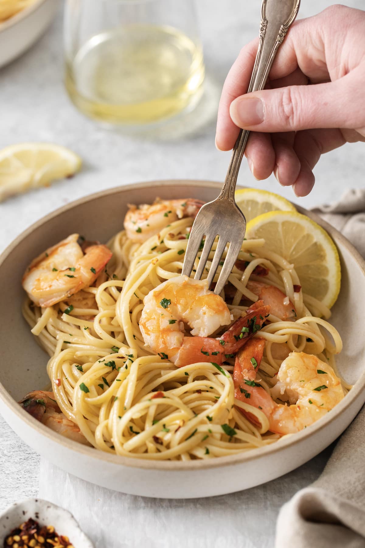 a hand holding a fork twirling shrimp pasta with linguine, red pepper flakes, herbs, and lemon slices in a bowl
