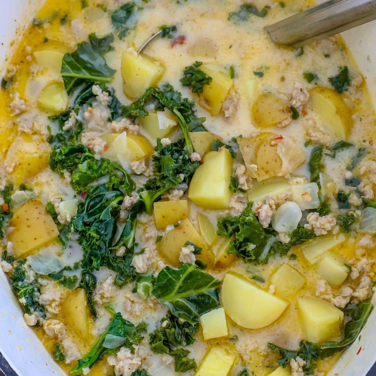 zuppa toscana soup with kale, potatoes, cream, and sausage in a white dutch oven with a ladle in it