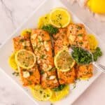 Three baked salmon fillets topped with lemon slices, garlic, and herbs on a white plate, garnished with parsley and lemon wedges. A spoon sprinkles more herbs on top—truly the best baked salmon recipe for any occasion.