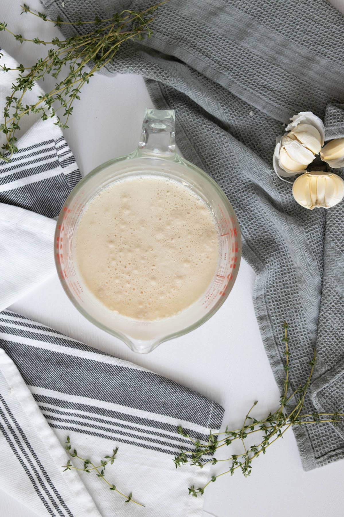 A measuring cup filled with garlic and herbs beside a table with overnight garlic herb focaccia.