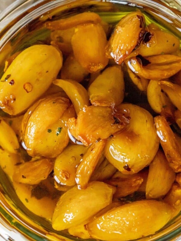 dark brown garlic confit in olive oil in a glass dish on a table next to a bulb of garlic