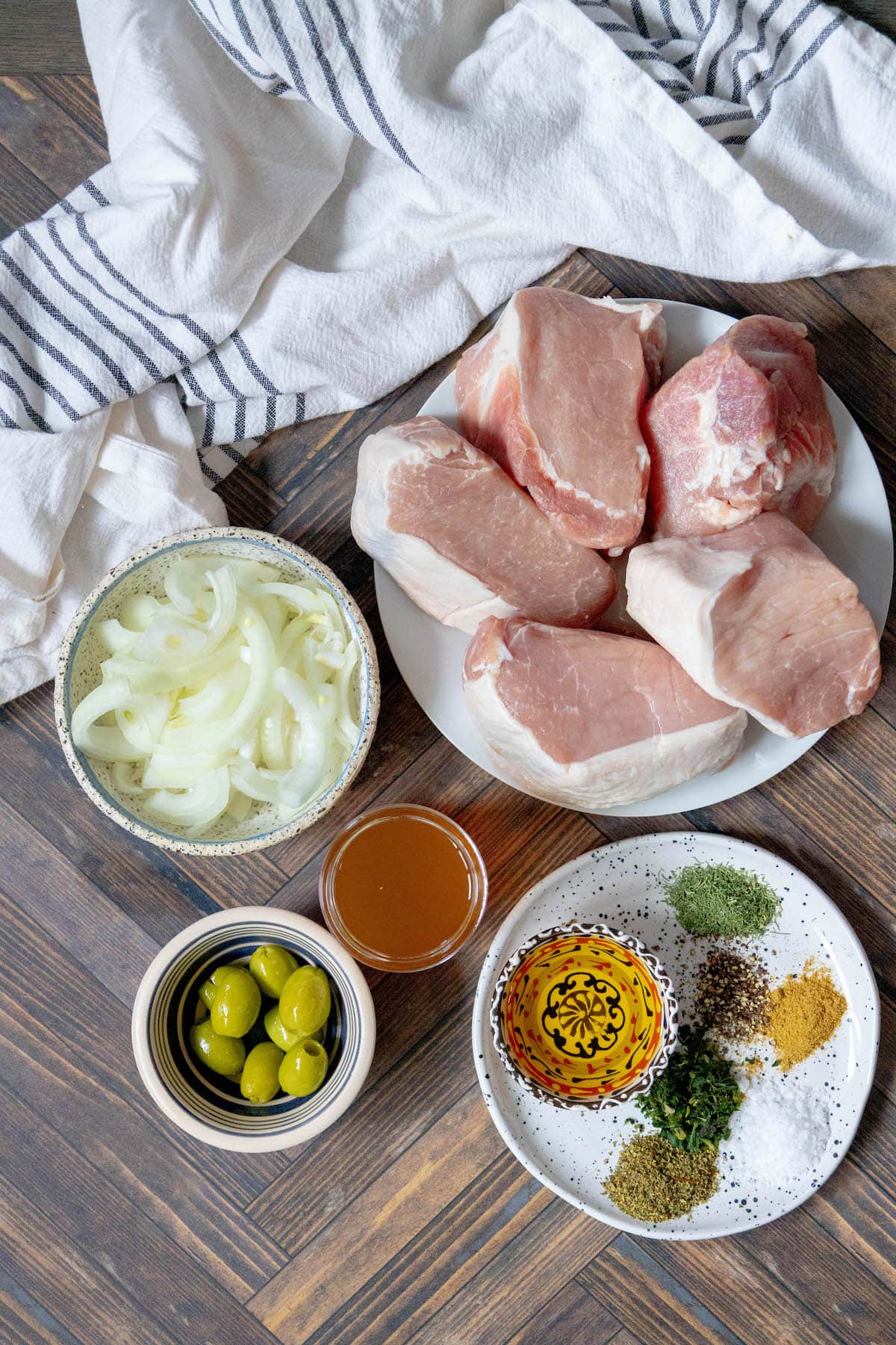 A collection of fresh ingredients for Greek-style pork chops, including herbs, olive oil, olives, onions, and stock, laid out on a rustic table.
