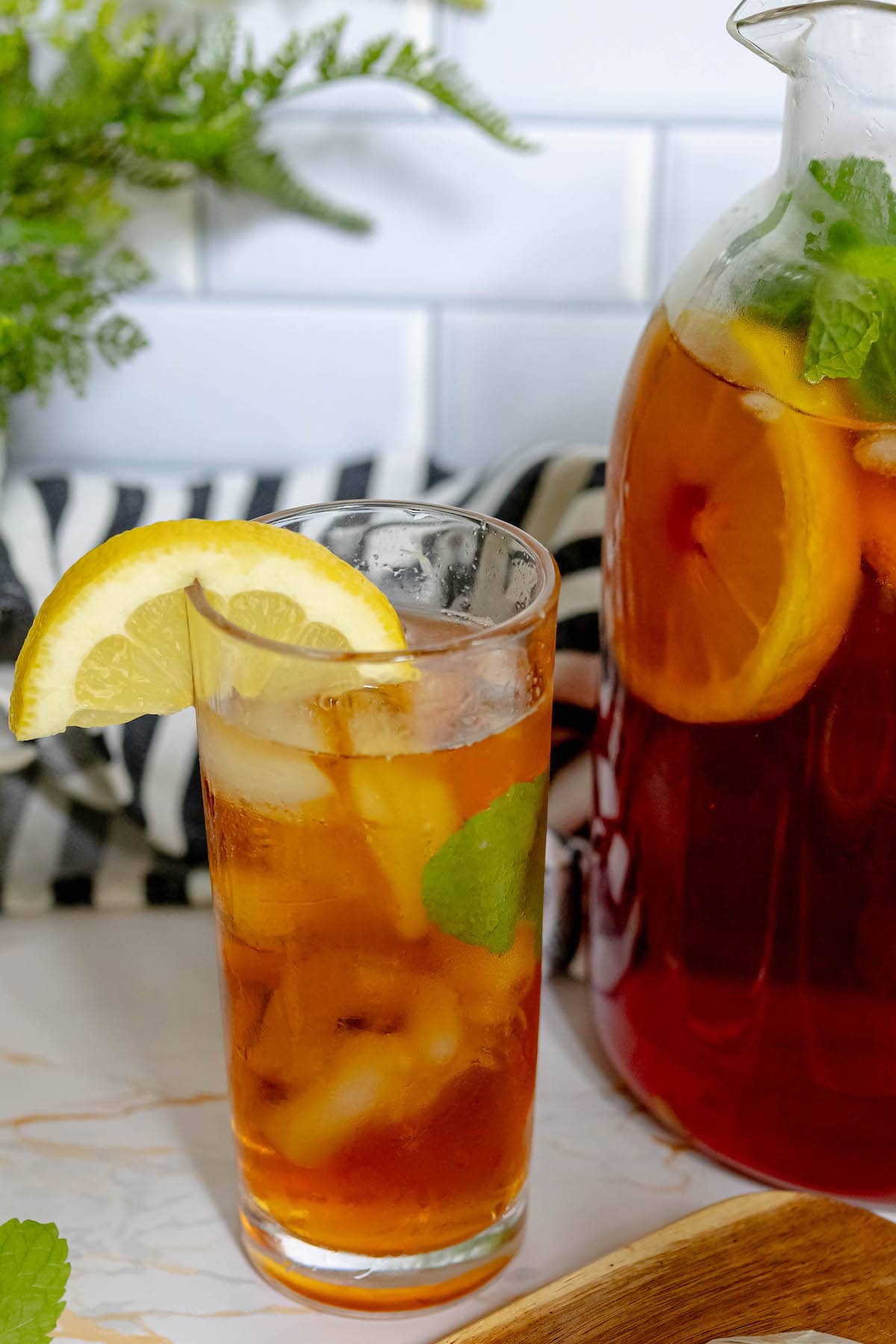 a tall glass of tea with a lemon slice and mint next to a pitcher of tea with lemon sliced and mint leaves in it 