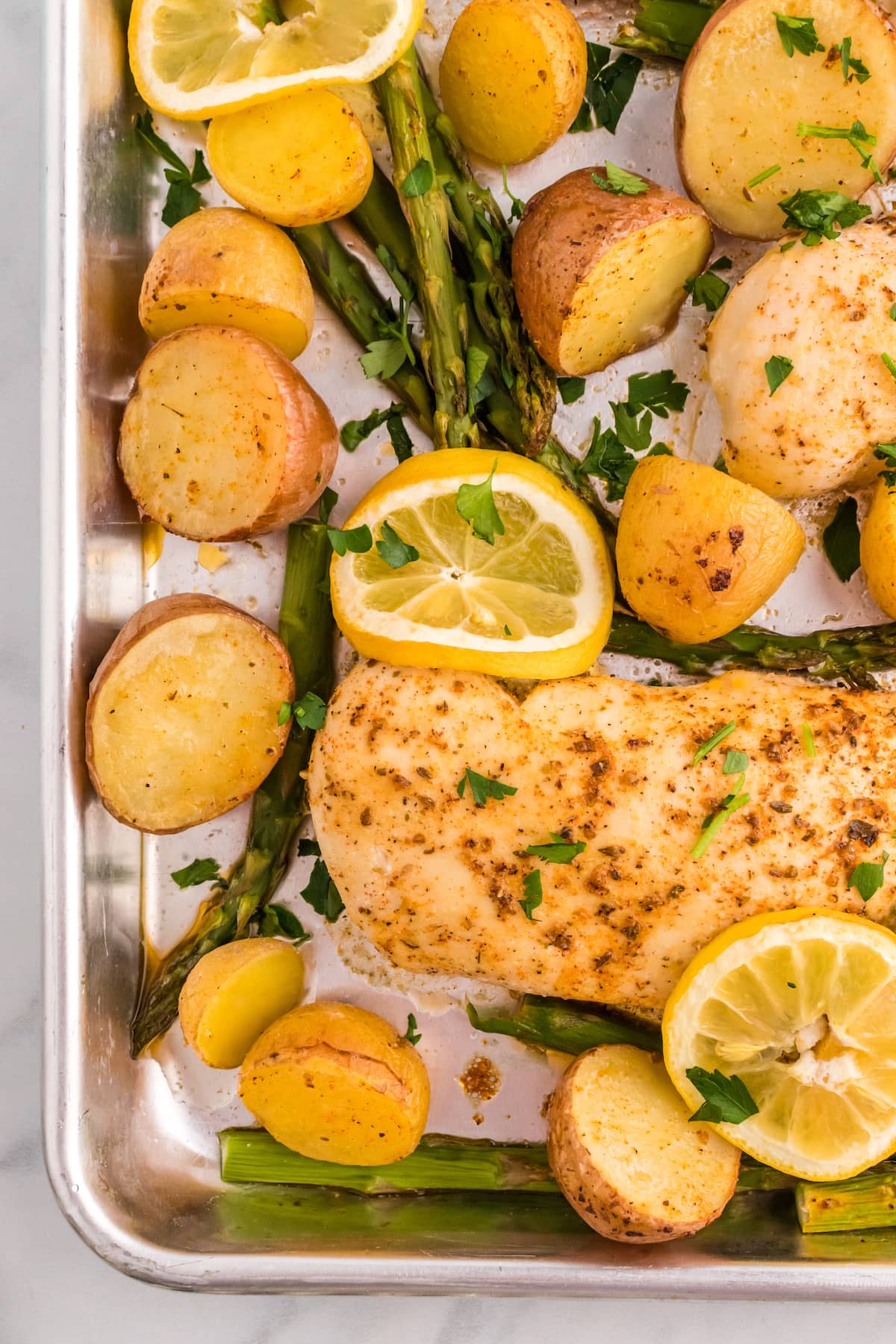 a sheet pan with baked chicken breasts, potatoes, lemon slices, and asparagus on it