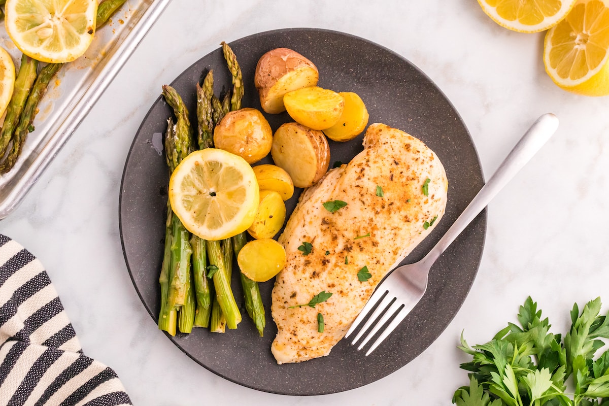 a black plate with baked chicken breasts, potatoes, lemon slices, and asparagus on it