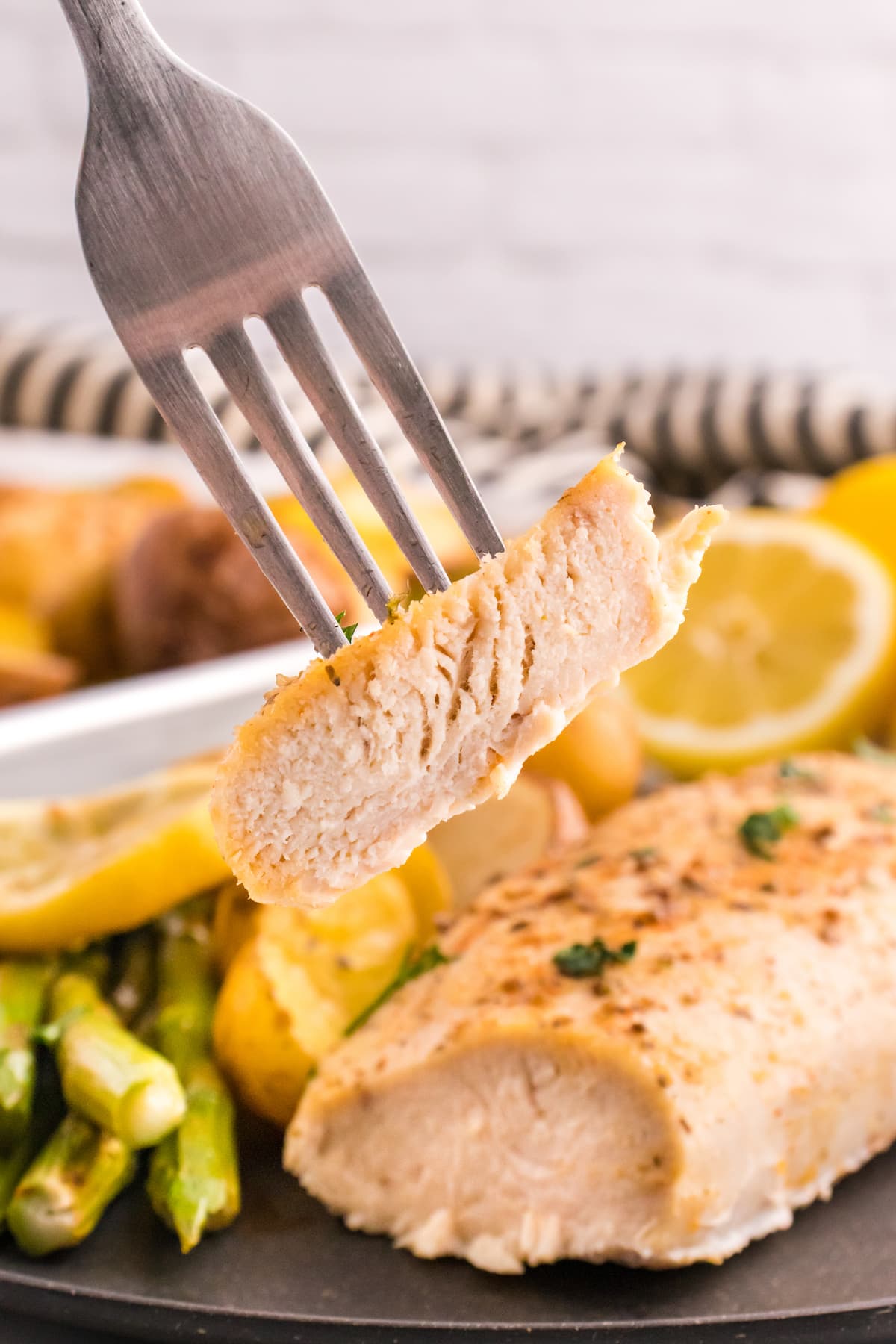 a fork with a piece of chicken on it over a plate with baked chicken breasts, potatoes, lemon slices, and asparagus on it