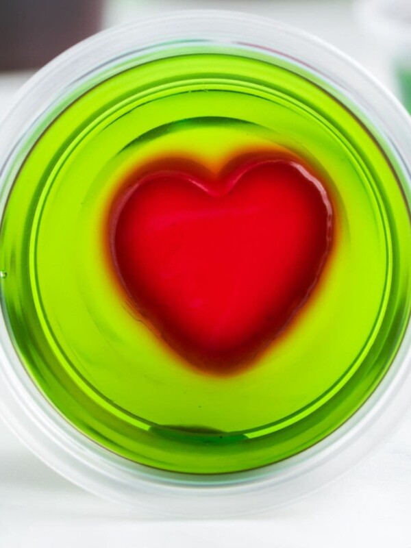 green jello shot with a red jello heart in the middle in a plastric cup on a table