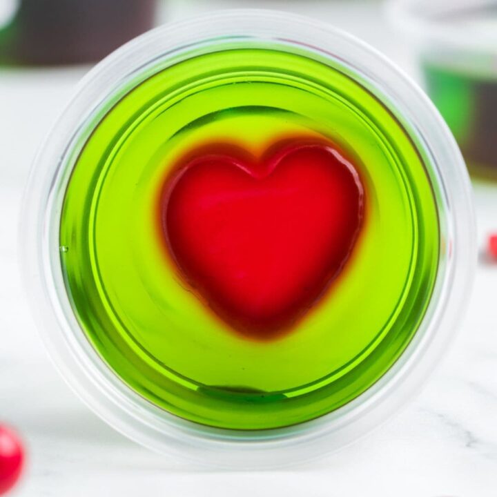 green jello shot with a red jello heart in the middle in a plastric cup on a table