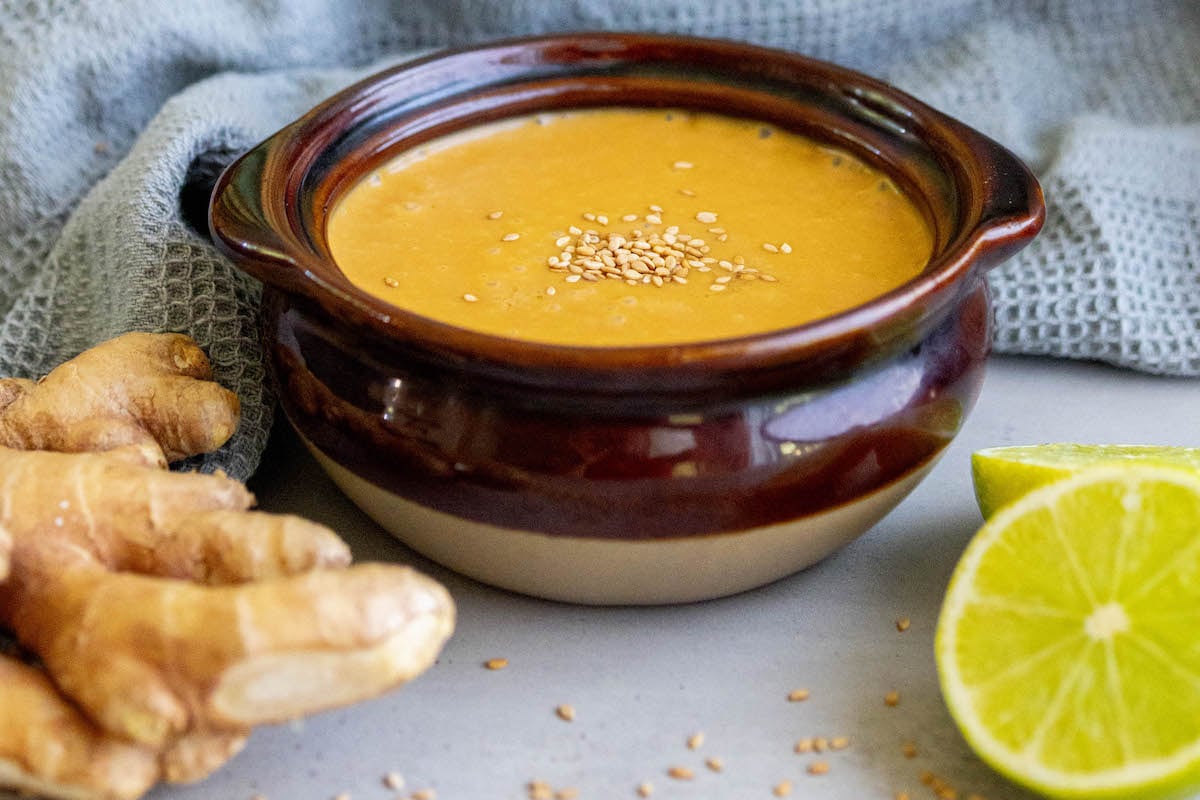 small brown pot of creamy dressing with sesame seeds with limes and ginger around it