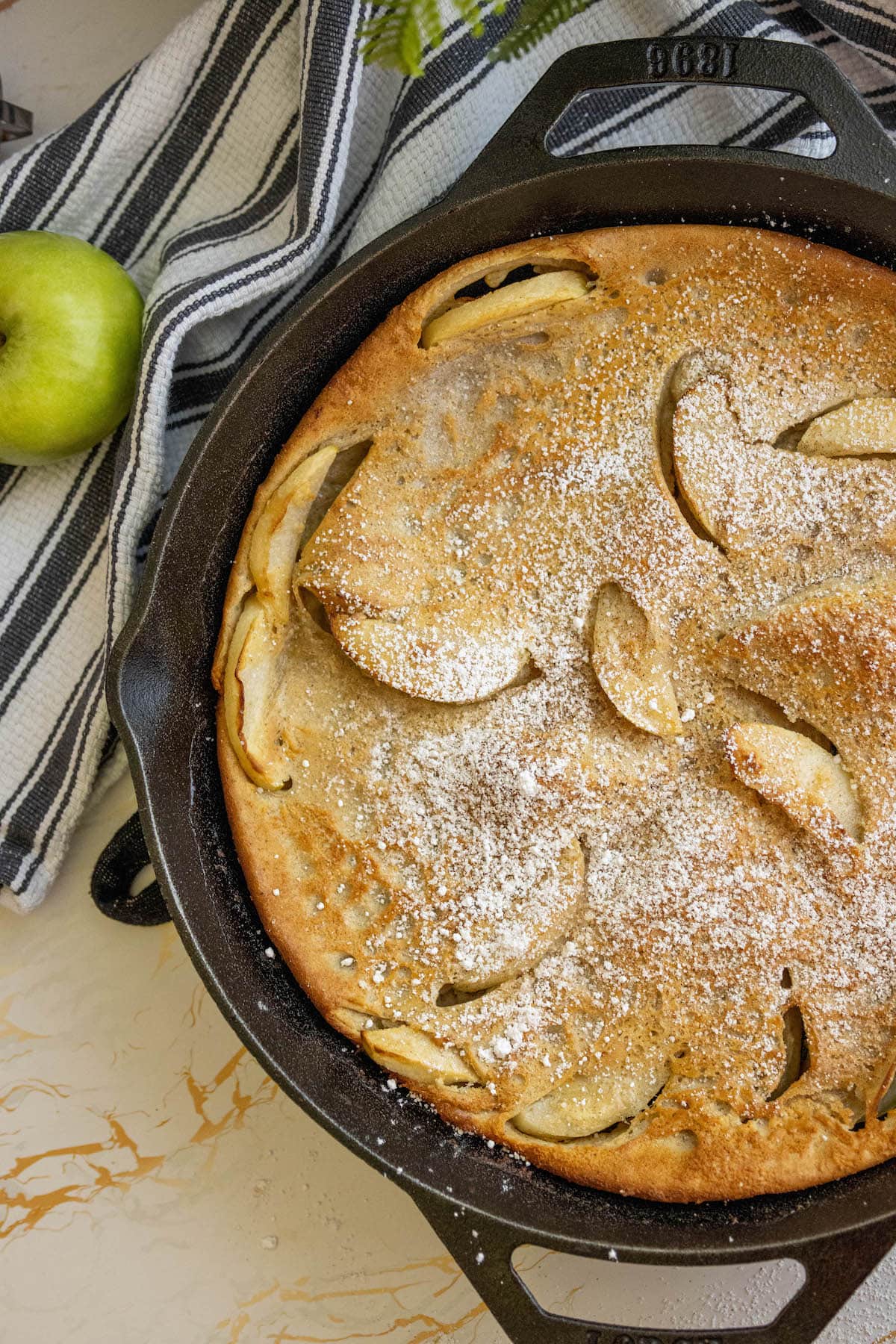 picture of a pancake with apples and powdered sugar on it in a cast iron skillet