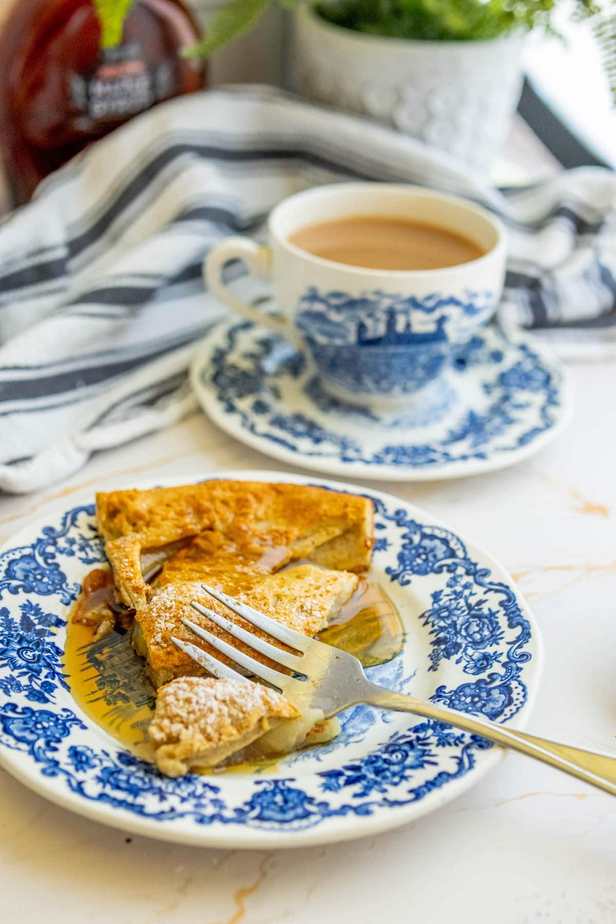 slice of popover pancake on a plate with powdered sugar and syrup, tea cup in the background