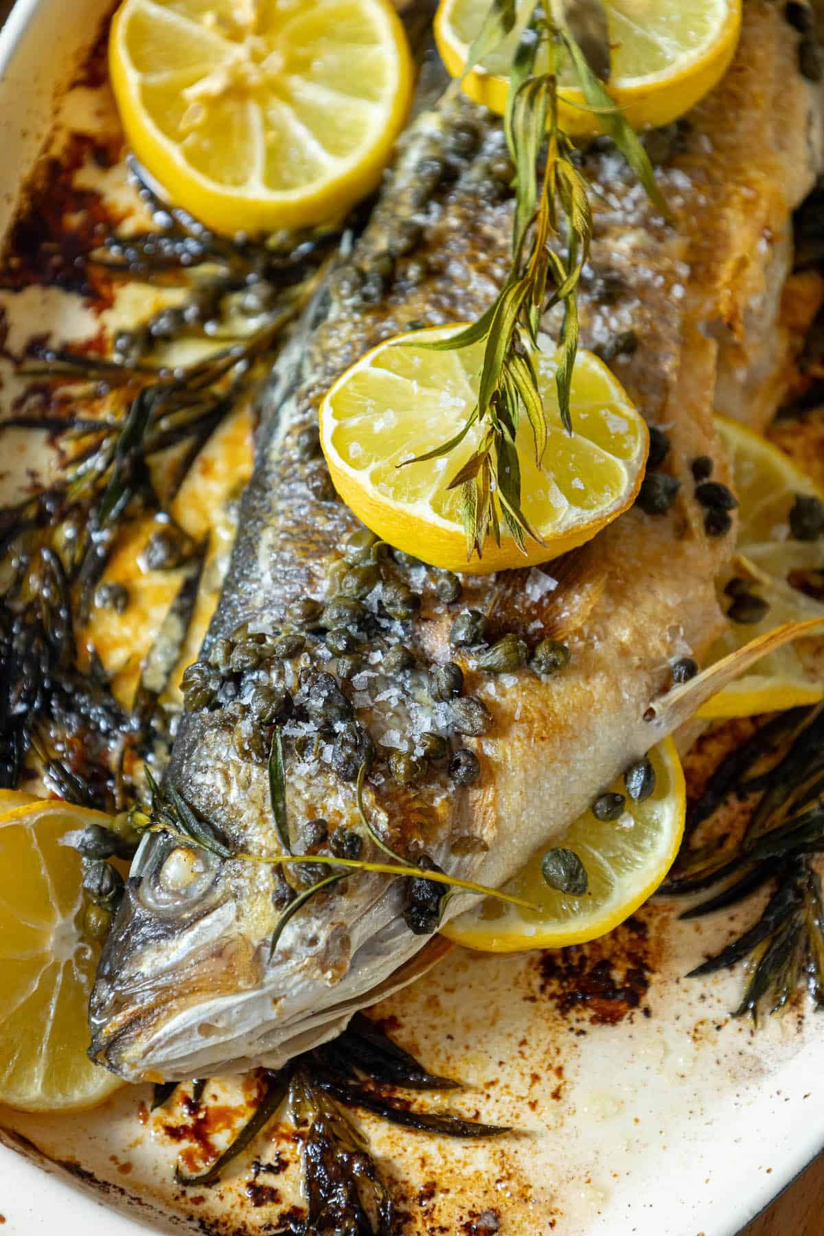 A lemon caper butter roasted branzino with herbs on a white plate.