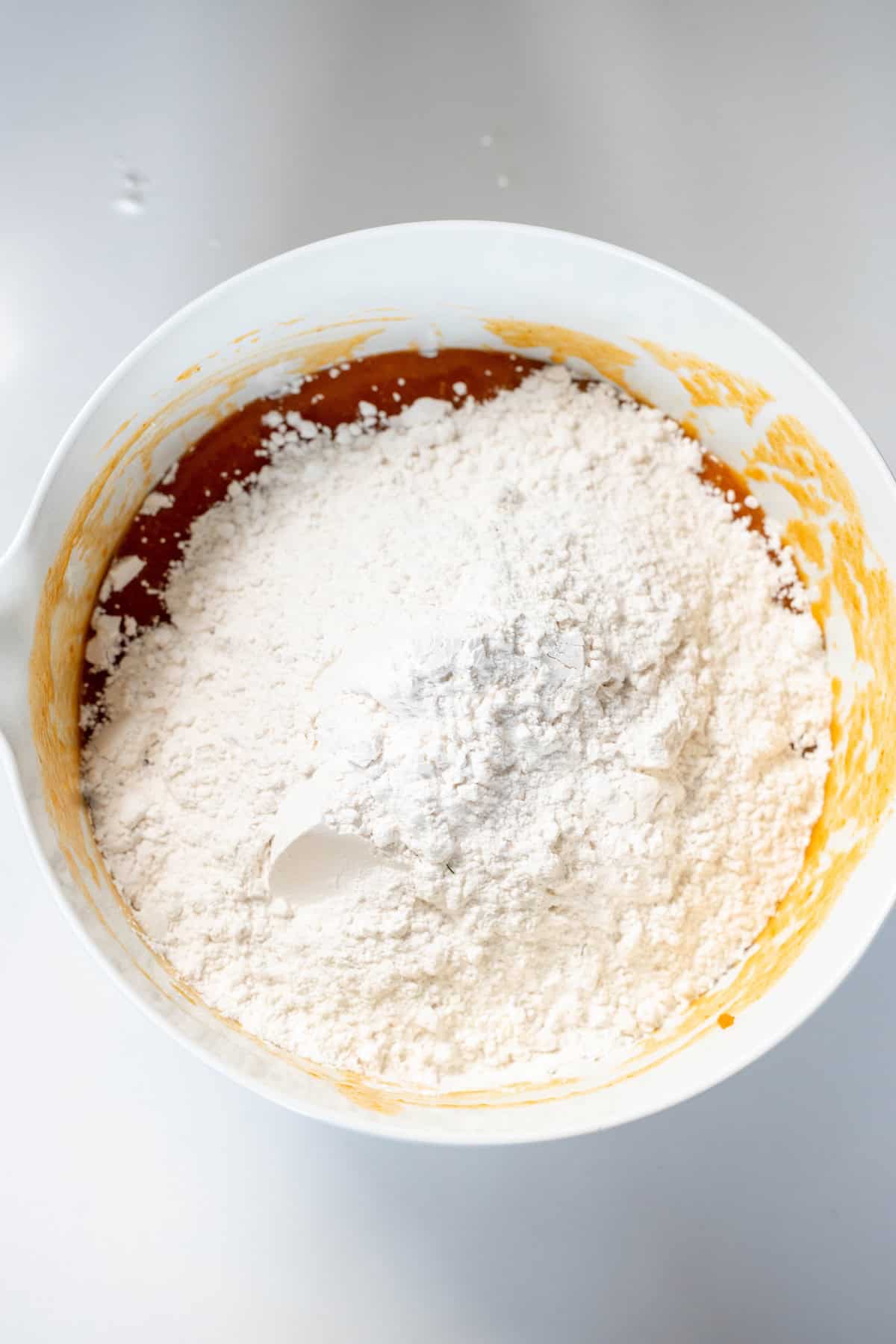 A white mixing bowl containing flour on top of a wet orange batter for a moist spiced pumpkin bread bundt cake recipe, with a white spatula partially buried in the mixture.