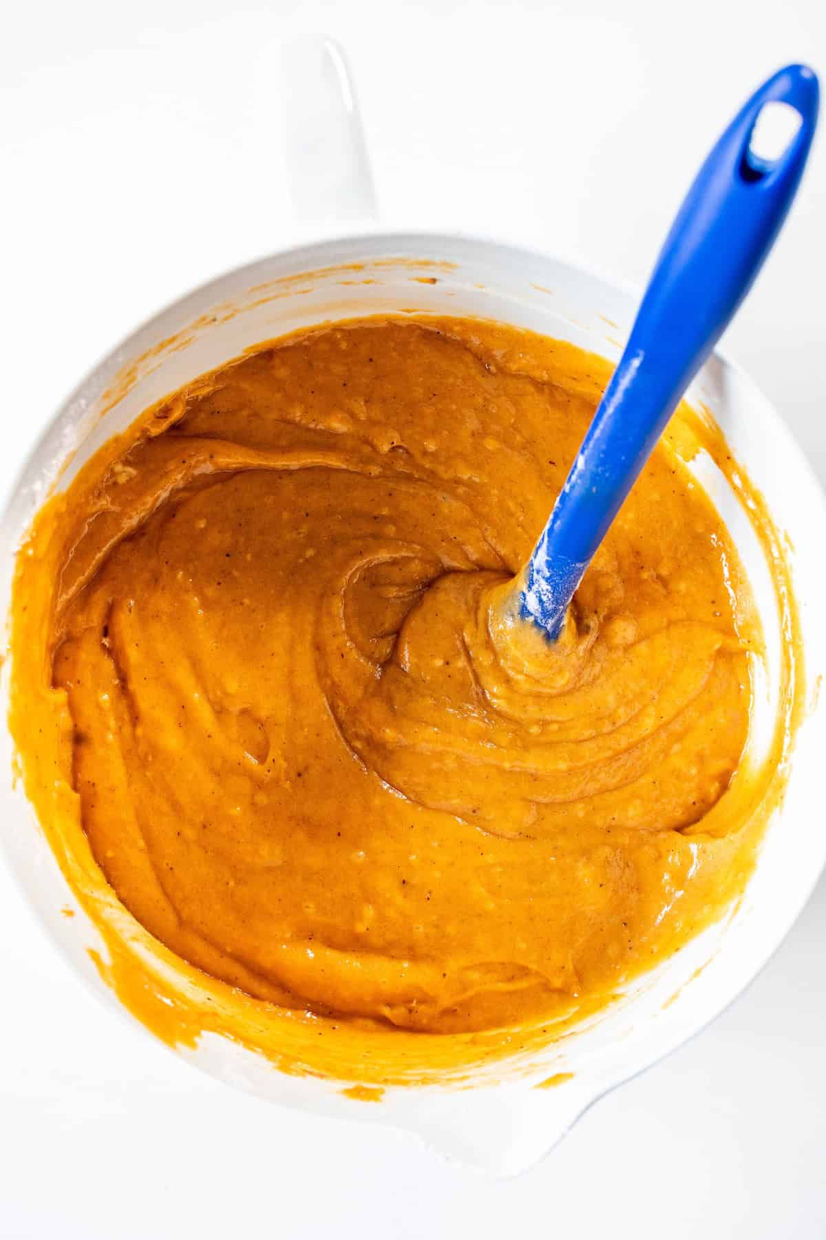 A bowl of orange batter being stirred with a blue spatula, viewed from above on a white surface—perfect for whipping up Halloween desserts like pumpkin spice bundt cake.