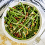 A bowl of cheesy sun-dried tomato green beans is mixed with pine nuts and crumbled cheese, sitting on a white surface with a striped towel and a fork nearby.