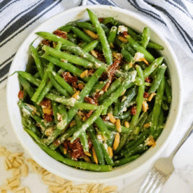 A bowl of cheesy sun-dried tomato green beans is mixed with pine nuts and crumbled cheese, sitting on a white surface with a striped towel and a fork nearby.