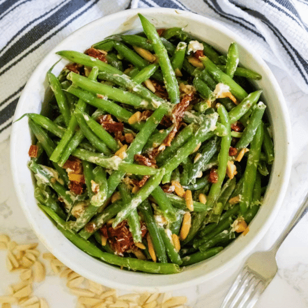 A bowl of cheesy sun-dried tomato green beans is mixed with pine nuts and crumbled cheese, sitting on a white surface with a striped towel and a fork nearby.